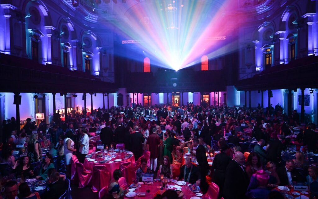 A large ballroom filled with circular tables of people in purple lighting
