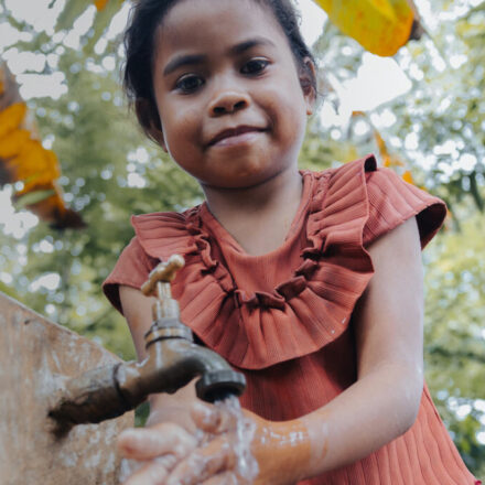 A young girl in a rust-coloured ruffled top washes her hands under a tap at a concrete water point, surrounded by lush green foliage. Overlay text reads 